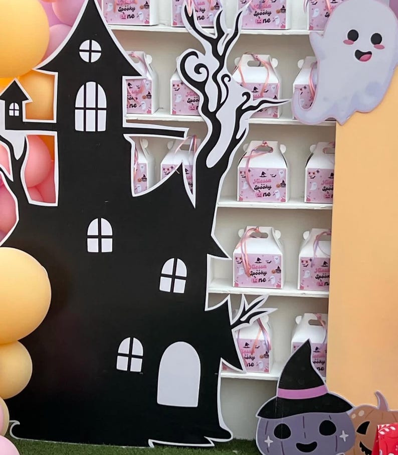 May include: A black haunted house cutout with white window details and a decorative tree. A shelf displays pink gift boxes with the text "Spooky One". A smiling ghost and a purple pumpkin with a witch hat are also present, creating a Halloween theme.
