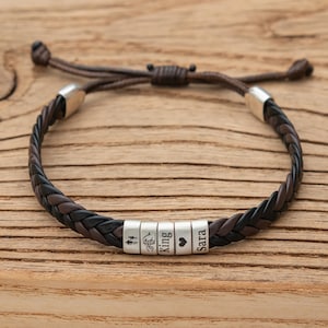 May include: Braided leather bracelet with brown and black strands. The bracelet features silver beads with engraved symbols and names. The adjustable cord is brown. The bracelet is displayed on a wooden surface.