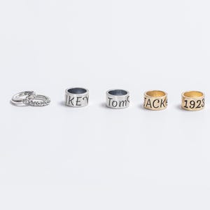 May include: A collection of five rings arranged in a row. The first is a silver-toned band with an intricate pattern. The next two are silver-toned bands engraved with the names Mikey and Tom. The final two are gold-toned bands engraved with JACK and 1923.