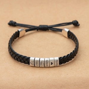 May include: A black braided leather bracelet featuring six cylindrical silver-toned beads. The beads are engraved with a line drawing of two people, the year 1985, a heart-shaped hand gesture, the name James, a heart symbol, and the name Sara.