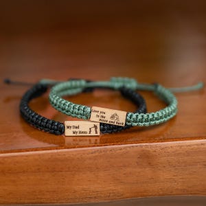 May include: Two braided cord bracelets featuring rectangular wooden charms. One bracelet is black with a charm engraved with "My Dad My Hero" and a silhouette. The other is olive green with a charm engraved with "Love you to the moon and back" and a silhouette.