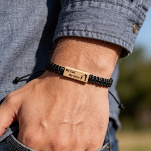 Op de afbeelding: Een zwarte armband van gevlochten koord met een rechthoekig houten middenstuk. Het hout is gegraveerd met de tekst My Dad My Hero en een silhouet van een volwassene die de hand van een kind vasthoudt. De armband heeft verstelbare uiteinden.