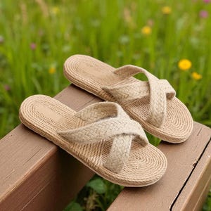 Op de afbeelding: Een paar beige geweven sandalen met kruisbanden, rustend op een houten oppervlak. De sandalen hebben een textuur die op touw lijkt en staan tegen een achtergrond van groen gras en wilde bloemen.