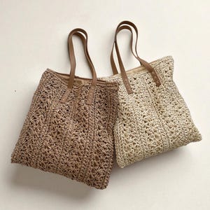 Puede incluir: Dos bolsos tote tejidos en beige y marrón. Los bolsos tienen forma rectangular con patrones calados y asas de cuero marrón. Los bolsos están diseñados para transportar objetos y tienen una estética casual y natural.