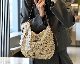 May include: A woven straw shoulder bag with a brown leather strap and a white lace bow. The bag has a rounded shape and a small brown leather patch. The bag is a light tan color.