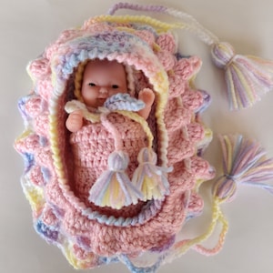 May include: A handmade crochet baby doll carrier in pastel colors. The carrier is pink with scalloped edges and a hood, and it has a small baby doll inside. The carrier has tassels and is made of soft yarn.