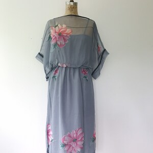 Op de afbeelding: Een grijze chiffonjurk met een bloemenprint. De jurk heeft een losse pasvorm en een getailleerde taille. De bloemen zijn roze en groen.