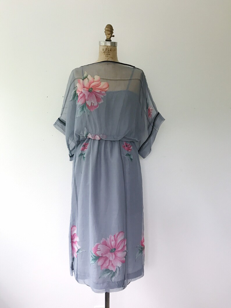 Op de afbeelding: Een grijze chiffonjurk met een bloemenprint. De jurk heeft een losse pasvorm en een getailleerde taille. De bloemen zijn roze en groen.