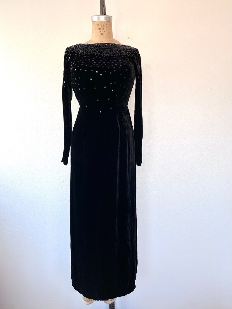 May include: A black velvet gown with a beaded neckline. The gown has long sleeves and a fitted bodice. The skirt is full and flows to the floor.