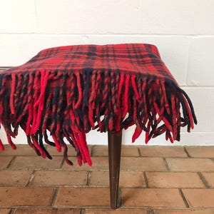 May include: Red and black plaid throw blanket with fringe. The blanket is draped over a wooden stool.