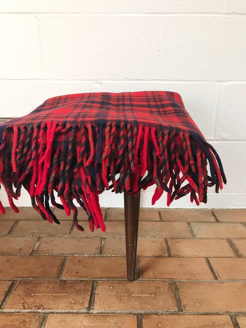 May include: Red and black plaid throw blanket with fringe. The blanket is draped over a wooden stool.
