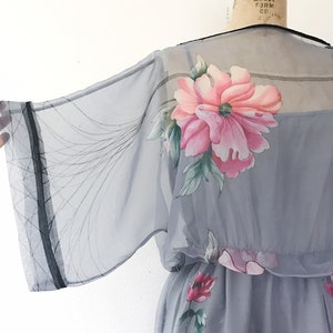 Op de afbeelding: Een grijze, doorschijnende jurk met een bloemenprint. De jurk heeft een getailleerde taille en een wijde, vloeiende mouw. De bloemen zijn roze, rood en groen.
