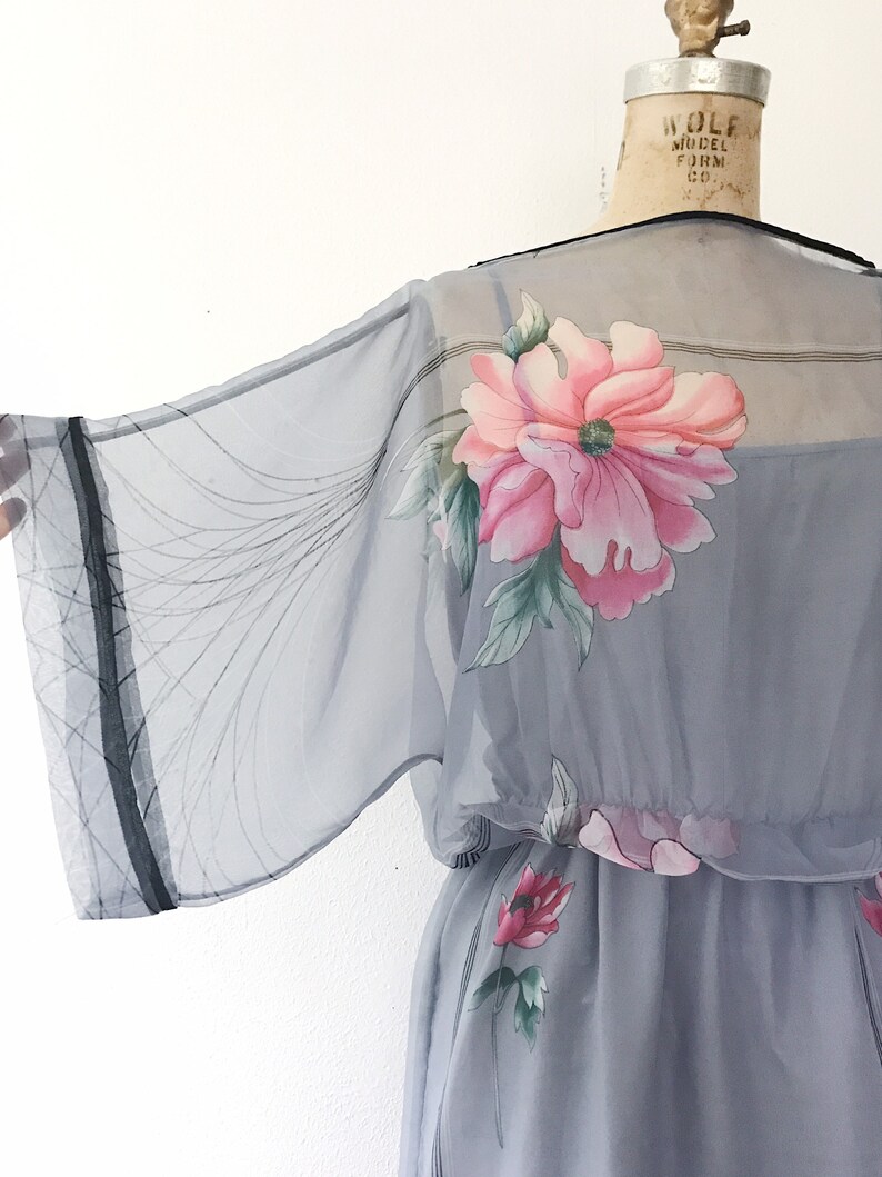 Op de afbeelding: Een grijze, doorschijnende jurk met een bloemenprint. De jurk heeft een getailleerde taille en een wijde, vloeiende mouw. De bloemen zijn roze, rood en groen.