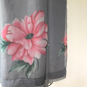 Op de afbeelding: Een grijze, doorschijnende jurk met een roze en groene bloemenprint. De jurk heeft een losse pasvorm en een zwierige silhouet.