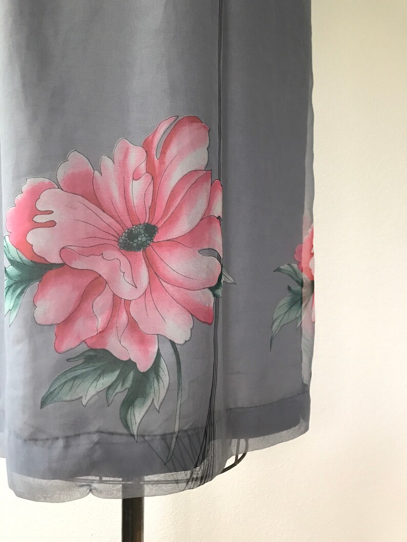 Op de afbeelding: Een grijze, doorschijnende jurk met een roze en groene bloemenprint. De jurk heeft een losse pasvorm en een zwierige silhouet.