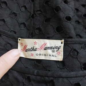 May include: A white label with black text that reads "Martha Manning Original" with a small flower design on each side of the text. The label is attached to a black fabric with a pattern of small circles.