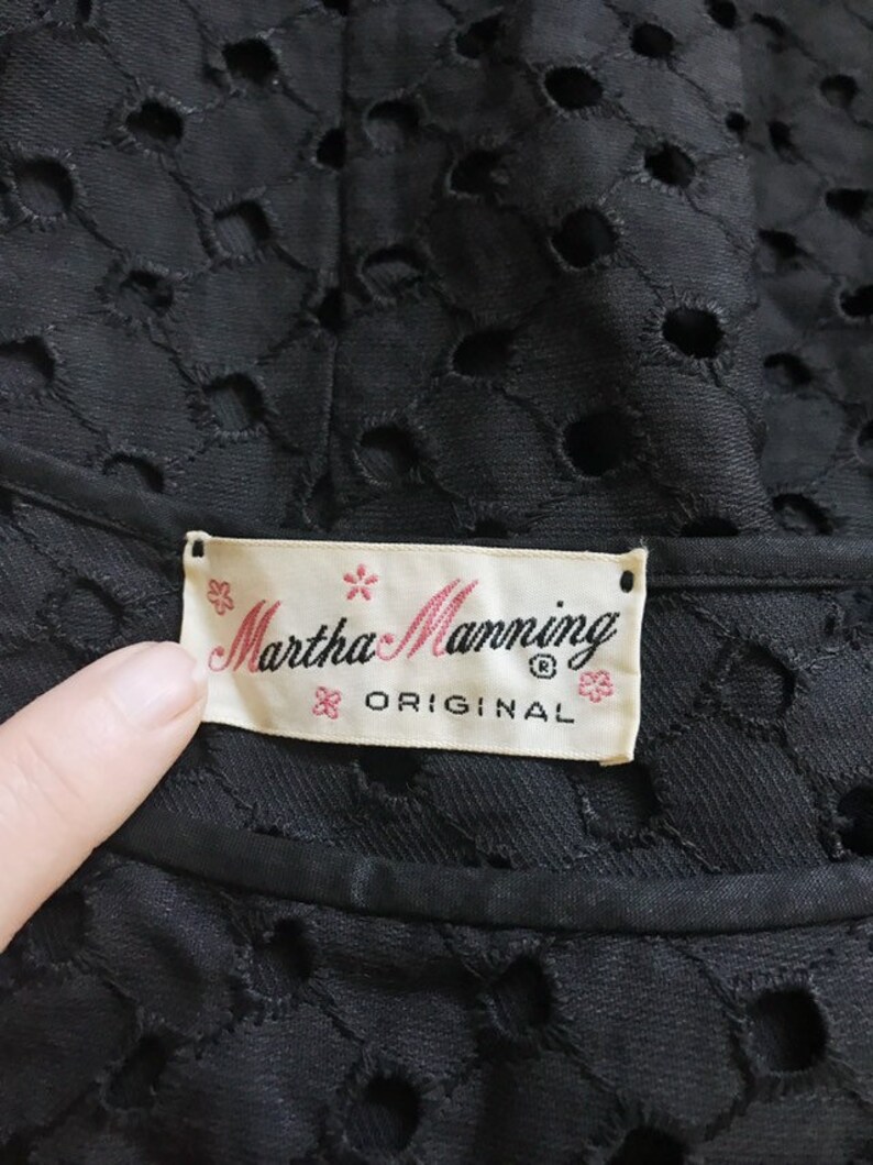 May include: A white label with black text that reads "Martha Manning Original" with a small flower design on each side of the text. The label is attached to a black fabric with a pattern of small circles.