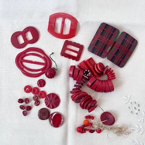 May include: A collection of vintage red buttons and buckles in various shapes and sizes. Includes round, square, and geometric designs. Some pieces have a glossy finish, while others are matte. Two plaid rectangular pieces are also present.