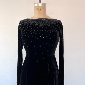 May include: A black velvet dress with a bateau neckline and long sleeves. The dress is embellished with black beads and rhinestones.