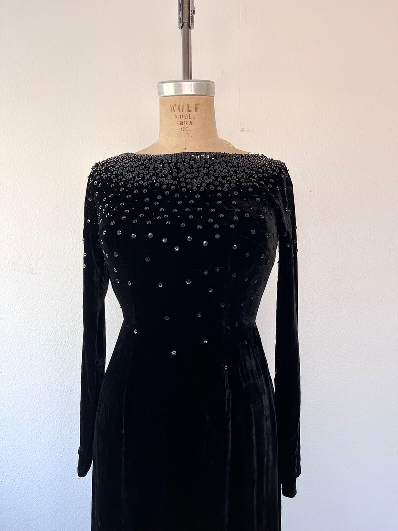May include: A black velvet dress with a bateau neckline and long sleeves. The dress is embellished with black beads and rhinestones.