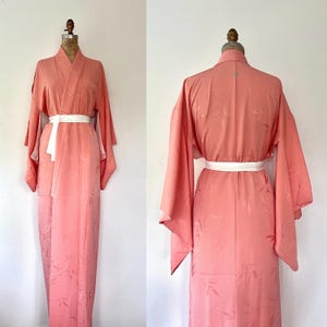 May include: A coral-colored kimono-style robe with wide sleeves and a white belt. The robe is displayed on a mannequin, showcasing its full length and design. The fabric has a subtle leaf pattern.