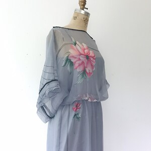 Op de afbeelding: Een lichtblauwe, doorschijnende jurk met een bloemenprint. De jurk heeft een getailleerde taille en een zwierige rok. De bloemenprint heeft roze en groene bloemen.