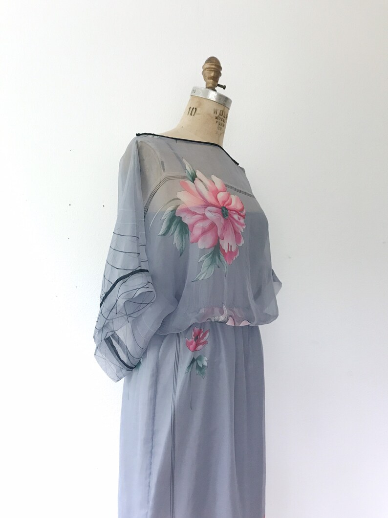 Op de afbeelding: Een lichtblauwe, doorschijnende jurk met een bloemenprint. De jurk heeft een getailleerde taille en een zwierige rok. De bloemenprint heeft roze en groene bloemen.