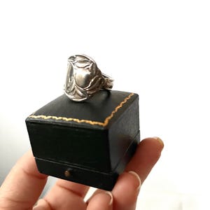 May include: A silver ring with a floral design sits atop a black jewelry box with gold trim. The ring's design features detailed leaves and curves. The box is held against a white background.