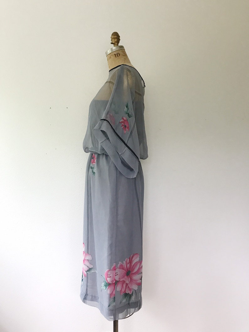 Op de afbeelding: Een grijze, doorschijnende jurk met een bloemenprint. De jurk heeft een losse pasvorm en een getailleerde taille. De bloemen zijn roze en groen.