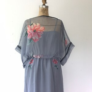 Op de afbeelding: Een grijze, doorschijnende jurk met een bloemenprint. De jurk heeft een getailleerd bovenstuk en een zwierige rok. De jurk is op een mannequin.