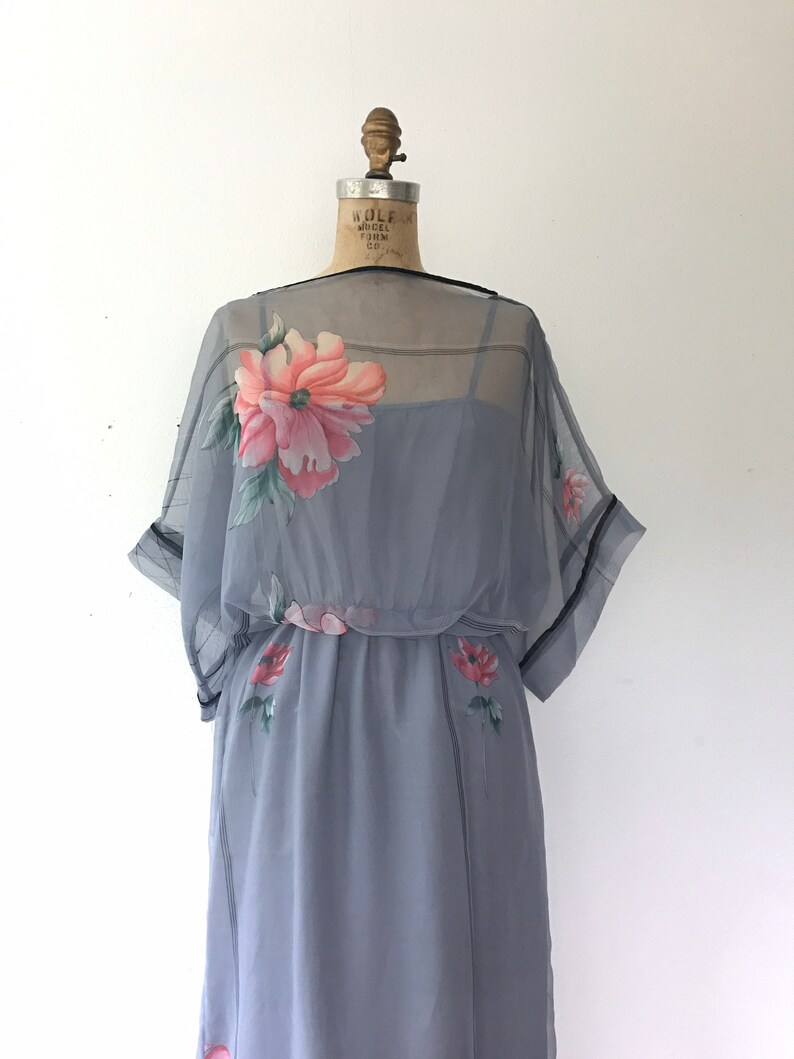 Op de afbeelding: Een grijze, doorschijnende jurk met een bloemenprint. De jurk heeft een getailleerd bovenstuk en een zwierige rok. De jurk is op een mannequin.