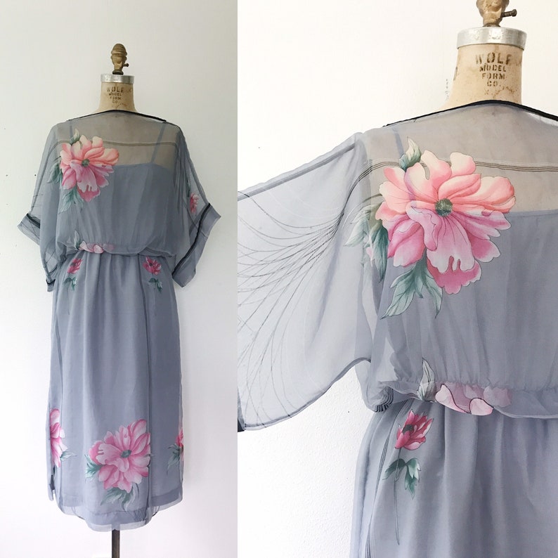 Op de afbeelding: Een grijze chiffonjurk met een bloemenprint. De jurk heeft een losse, vloeiende silhouet en een getailleerde taille. De mouwen zijn kort en hebben een klokvorm. De jurk is versierd met roze en groene bloemen.
