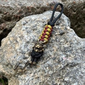 May include: A keychain featuring a braided paracord lanyard in alternating gold and maroon colors, topped with a black loop. At the base, a metallic bronze-toned samurai helmet charm sits above a black knotted cord with two small cylindrical beads.