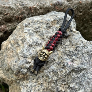 May include: A keyring featuring a brass-coloured Hannya mask bead attached to a black and red braided paracord lanyard. The cord forms a loop at the top and ends with two knotted black cord tails, each finished with a small metallic bead at the base.