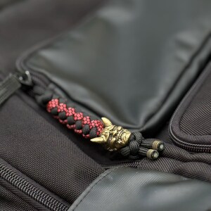 May include: A black and red braided paracord zip pull featuring a metallic brass-toned Hannya mask bead with horns. The accessory is attached to the zip of a black textured fabric bag, showcasing intricate knotting and a detailed face design.