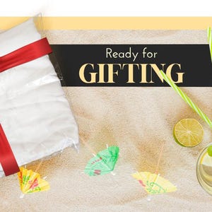 Pu&ograve; includere: Una borsa regalo bianca con un nastro e un fiocco rosso, pronta per essere regalata. La borsa &egrave; su una superficie sabbiosa con piccoli ombrellini decorativi e un bicchiere di limonata.