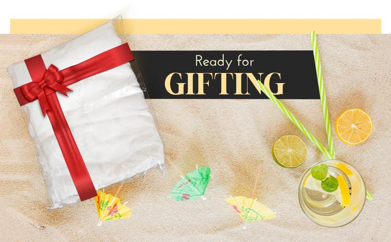 Pu&ograve; includere: Una borsa regalo bianca con un nastro e un fiocco rosso, pronta per essere regalata. La borsa &egrave; su una superficie sabbiosa con piccoli ombrellini decorativi e un bicchiere di limonata.