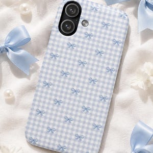 Pode incluir: Uma capa retangular para smartphone com um padrão de xadrez vichy em azul claro e branco, decorada com pequenos ícones de laços azul claro repetidos. A capa inclui duas aberturas circulares para as lentes da câmara e uma para o flash.