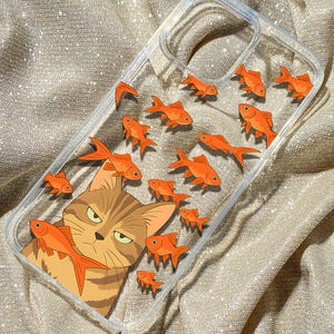 May include: A clear phone case with a cartoon cat and orange goldfish printed on it. The case is on a sparkly gold fabric.