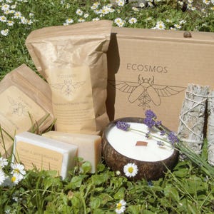 May include: A collection of natural wellness items, including a coconut shell candle, sage bundles, soaps, and packaged goods, set on grass and daisies. The products are branded with the "ECOSMOS" logo.