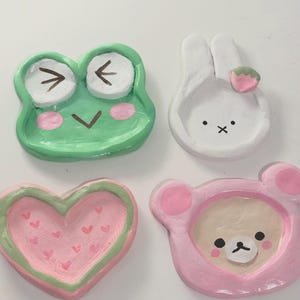 Puede incluir: Cuatro pequeños platos de cerámica hechos a mano. Una rana verde, un conejito blanco con una fresa, un corazón rosa con pequeños corazones y un oso rosa. Cada plato está pintado a mano.
