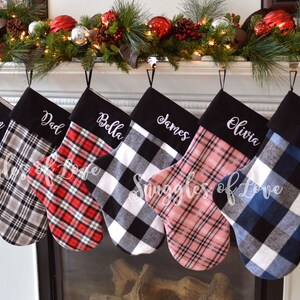 May include: Six personalized Christmas stockings with black and white plaid fabric. The stockings are hung on a fireplace mantel and are embroidered with the names "Mom", "Dad", "Bella", "James", "Olivia", and "Noah".