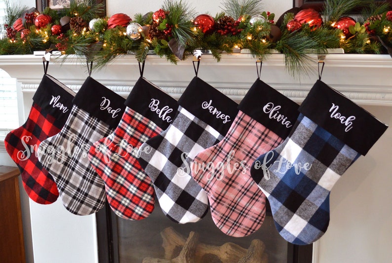 May include: Six personalized Christmas stockings with black and white plaid fabric. The stockings are hung on a fireplace mantel and are embroidered with the names "Mom", "Dad", "Bella", "James", "Olivia", and "Noah".