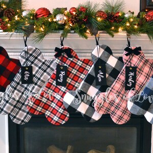 May include: Six personalized dog bone-shaped Christmas stockings with plaid patterns in red, black, white, blue, and pink. The stockings are hung on a white fireplace mantel with a garland of greenery and lights. The stockings are labeled with the names Millie, Elvis, Rexy, Alex, Harley, and  Baxter.
