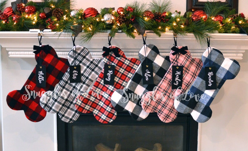 May include: Six personalized dog bone-shaped Christmas stockings with plaid patterns in red, black, white, blue, and pink. The stockings are hung on a white fireplace mantel with a garland of greenery and lights. The stockings are labeled with the names Millie, Elvis, Rexy, Alex, Harley, and  Baxter.