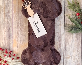 May include: A brown, bone-shaped Christmas stocking with a plush texture. A wooden deer ornament and a tag with the name "Storm" are attached. Festive greenery and red berries are in the background.