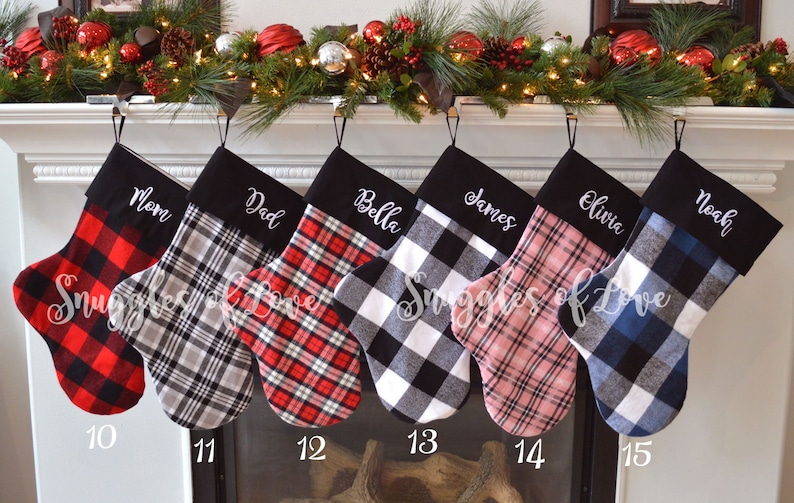 May include: Six personalized Christmas stockings with black felt cuffs and plaid fabric. The stockings are hung on a fireplace mantel with a garland of greenery and red ornaments. The names on the stockings are Mom, Dad, Bella, James, Olivia, and Noah.