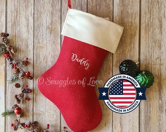 Puede incluir: Calcetín de Navidad de fieltro rojo con un puño de piel sintética blanca, bordado con la palabra "Daddy" y un logotipo blanco "Snuggles of Love". Una bandera estadounidense roja, blanca y azul está en un círculo con el texto "Handmade in America" sobre un fondo blanco.