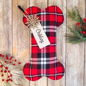 May include: A dog bone-shaped Christmas stocking in red, black, and white plaid. A tag with the name "Oakley" hangs from a gold snowflake ornament. The stocking has a black loop for hanging and is decorated with festive greenery.