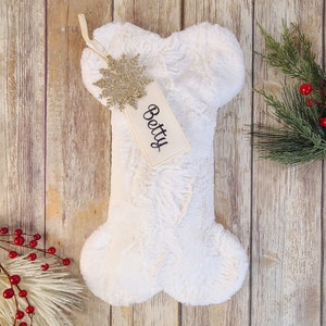 May include: White plush bone-shaped stocking with a gold snowflake ornament and a white tag that says "Betty".